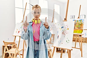 Young caucasian girl at art studio pointing up looking sad and upset, indicating direction with fingers, unhappy and depressed