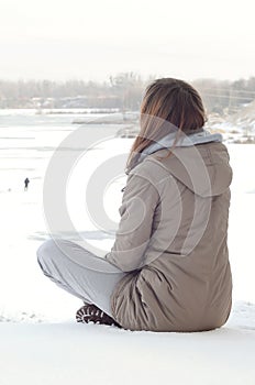 A young Caucasian girl in a brown coat staring into the distance on the horizon line between the sky and the frozen lake in winter