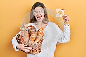 Young caucasian girl holding wicker basket with bread and loaf with heart shape smiling and laughing hard out loud because funny
