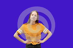 Young caucasian girl with long brown hair thinks, holds her hands on her belt, looks away, isolated on blue background