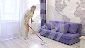 A young Caucasian girl moves around the house on one crutch and cleans the floors with a mop with one hand. Leg injury