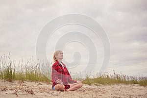 A young Caucasian girl in a red shirt and blue denim shorts sits on the sand by the river