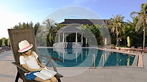 Young caucasian girl sitting on armchair and reading book. Swimming pool and palm trees at background. Have same clip