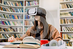 Young Caucasian girl student in brown shirt sitting at the table with books in college library and studying using VR