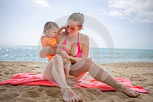 Young caucasian girl in a swimsuit and glasses sits on the sand at the beach,holds a cute baby in her arms and