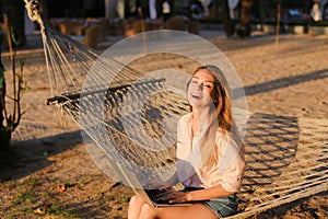 Young caucasian girl using laptop and sitting on sand in wicker hammock.