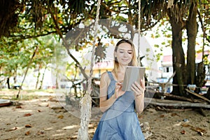 Young caucasian girl using tablet and riding swing on sand, wearing jeans sundress.