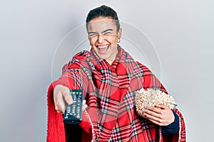 Young caucasian girl wearing blanket eating popcorn using tv control smiling and laughing hard out loud because funny crazy joke