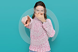 Young caucasian girl wearing wool winter sweater laughing at you, pointing finger to the camera with hand over mouth, shame