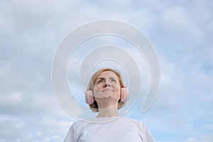 A young Caucasian girl in a white T-shirt and headphones stands against the background of a blue sky with clouds.