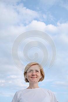 A young Caucasian girl in a white T-shirt and headphones stands against the background of a blue sky with clouds.