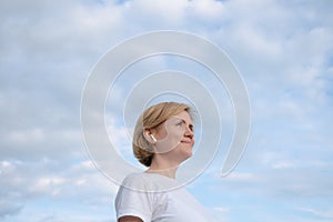 A young Caucasian girl in a white T-shirt and headphones stands against the background of a blue sky with clouds.