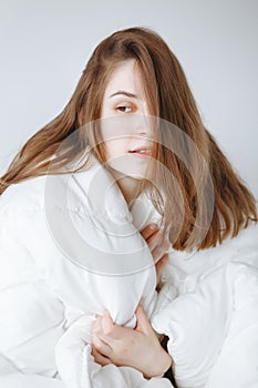 Young Caucasian girl woman with long red hair sitting in bed wrapped covered with white blanket