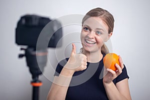 Young caucasian girl woman making a video blog vlog with camera and orange about healthy vegetarian lifestyle