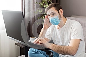 Young caucasian gloomy man in stress working from home office wearing protective mask using laptop and internet. Coronavirus covid