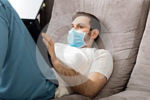 Young caucasian gloomy man working from home office wearing protective mask using laptop and internet. Coronavirus covid 19