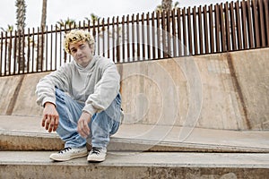 Young caucasian guy with blonde hair sitting on street and looking at camera.