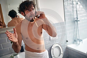 Young caucasian guy looking confused and horrified  after receiving bad news on his cell phone while brushing his teeth in the photo