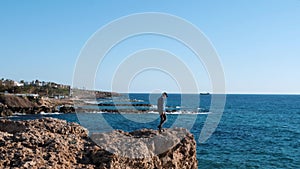 Young caucasian guy runs on rocks to edge of cliff. Boy in blue shirt and jeans raises hands to sky. Guy running to edge of cliff