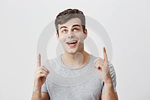 Young caucasian guy wearing gray t-shirt pointing at copy space up with forefingers looking with broad smile upwards