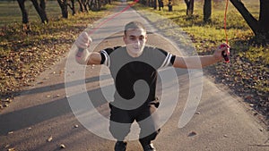 Young Caucasian handsome man in black sportswear jumping squats with jump rope in the autumn park. Athletic boy training