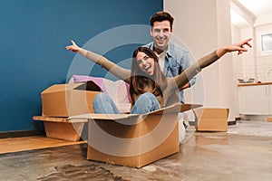 Young caucasian happy couple having fun playing with cardboard box in their new house after moving. Husband and wife
