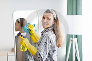 Young Caucasian housewife cleaning mirror, using spray detergent and rag at home. Sanitary service concept