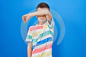 Young caucasian kid standing over blue background covering eyes with arm, looking serious and sad