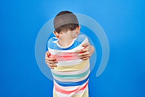 Young caucasian kid standing over blue background hugging oneself happy and positive from backwards
