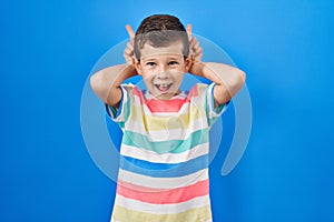 Young caucasian kid standing over blue background posing funny and crazy with fingers on head as bunny ears, smiling cheerful