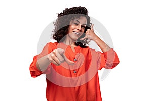 young caucasian lady with black curly hair dressed in an orange blouse points her finger at the camera