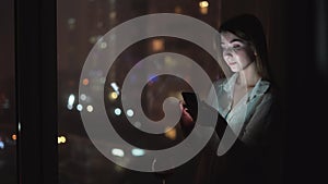 A young caucasian lady stands at the window at night, chatting with friends using her smartphone. Lights of the night