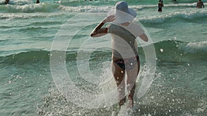 Young Caucasian Lady Woman In Swimsuit And Summer Hat Running In Sea Water Splashes. Vacation On Sea Ocean Beach. slow