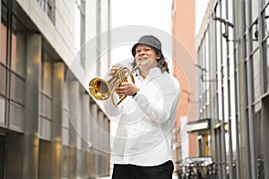 Young caucasian long-haired blond man in shirt and hat playing trumpet outdoor