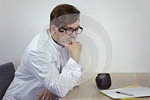 Young caucasian male doctor in a white outfit sits thoughtful, holding his chin, with cup of tea or coffee and papers on the table