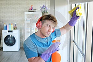 Young Caucasian male Wearing violet rubber gloves is holding Cleanser bottle and sponge. He is cleaning and wiping on glass window
