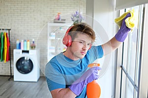 Young Caucasian male Wearing violet rubber gloves is holding Cleanser bottle and sponge. He is cleaning and wiping on glass window