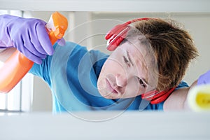 Young Caucasian male Wearing violet rubber gloves is holding Cleanser bottle and sponge. He is cleaning and wiping on white Shelf