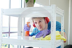 Young Caucasian male Wearing violet rubber gloves is holding Cleanser bottle and sponge. He is cleaning and wiping on white Shelf