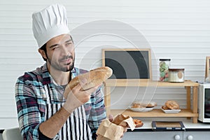 Young Caucasian man with beard wear chef hat and apron holding loaf of bread with pride and looking forward with a determined eye