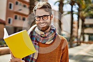 Young caucasian man with beard wearing glasses reading a book outdoors on a sunny day