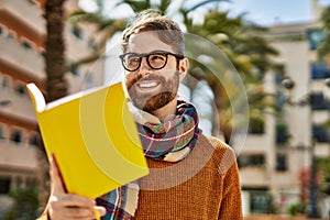 Young caucasian man with beard wearing glasses reading a book outdoors on a sunny day