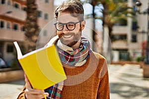 Young caucasian man with beard wearing glasses reading a book outdoors on a sunny day