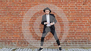 A young caucasian man in a black suit and hat is dancing and fooling around against a brick wall, slow-mo, copy space
