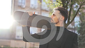 Young Caucasian man in black suit looking away holding skateboard on shoulder. Medium shot portrait of smiling handsome