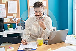 Young caucasian man business worker talking on smartphone writing on paperwork at office