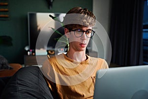 Young caucasian man in casual clothing and eyeglasses using laptop in living room on sofa at home