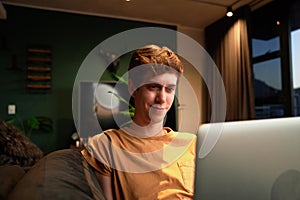 Young caucasian man in casual clothing smiling while using laptop on sofa at home