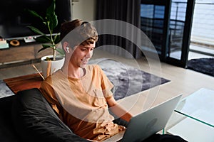 Young caucasian man in casual clothing using laptop with wireless headphones on sofa at home