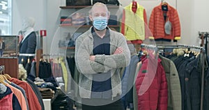 Young Caucasian man in Covid-19 face mask crossing hands and looking at camera as blurred woman choosing clothes at the